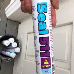 ELECTRO CEL Instalacje elektryczne - Pojemnik z masą uszczelniającą Seal 511 trzymany w dłoni na tle białych, frezowanych drzwi w Warszawie. Obok wiaderko z zapasowymi tubami uszczelniacza.