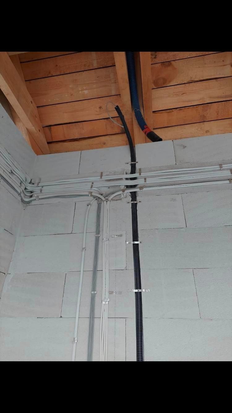 Białe i czarne kable elektryczne prowadzone po ścianie z bloczków betonowych pod drewnianym stropem, widoczne metalowe uchwyty mocujące.