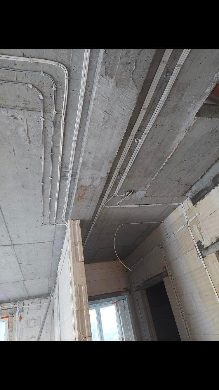 Szare, surowe wnętrze w trakcie budowy z widocznymi białymi, plastikowymi rurami i listwami elektrycznymi zamontowanymi na suficie i ścianach.