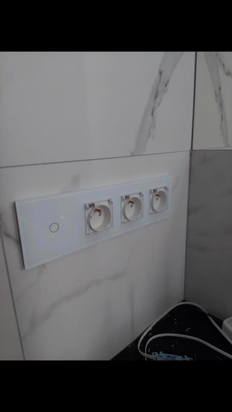 Potrójne gniazdko elektryczne z białym szklanym panelem na marmurowej ścianie, widoczne przewody w dolnej części kadru.