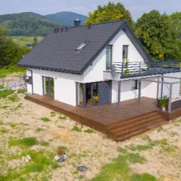 Nowoczesny dom z białymi ścianami i ciemnym dachem, z dużym tarasem z drewna i pergolą, widok z góry na tle zielonych wzgórz.