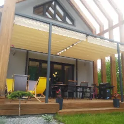 Zadaszony taras z drewnianą podłogą, meblami ogrodowymi i grillem, osłonięty beżową, zwijaną markizą, z widoczną konstrukcją drewnianą i zielenią w tle.