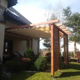 Drewniana konstrukcja z beżowym, falistym zadaszeniem przylegająca do domu, z widocznymi elementami ażurowej ścianki bocznej, na tle zielonego trawnika i iglastego drzewa.
