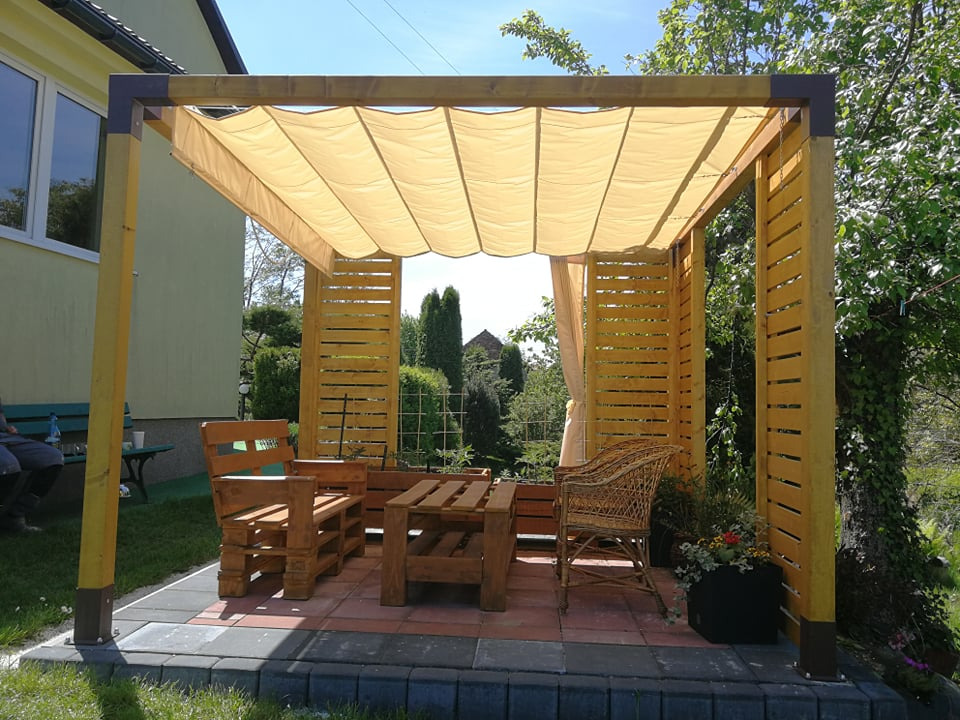 Drewniana konstrukcja z beżowym, falującym zadaszeniem materiałowym, tworząca zacienione miejsce do siedzenia z meblami z palet i wiklinowym fotelem, na wyłożonym płytkami tarasie w ogrodzie.