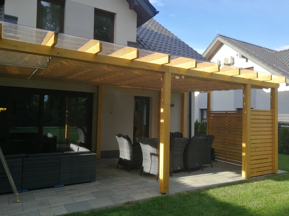 Zadaszona konstrukcja z jasnego drewna z przezroczystym dachem, przylegająca do domu, z widocznym zestawem mebli ogrodowych i fragmentem drewnianego ekranu.