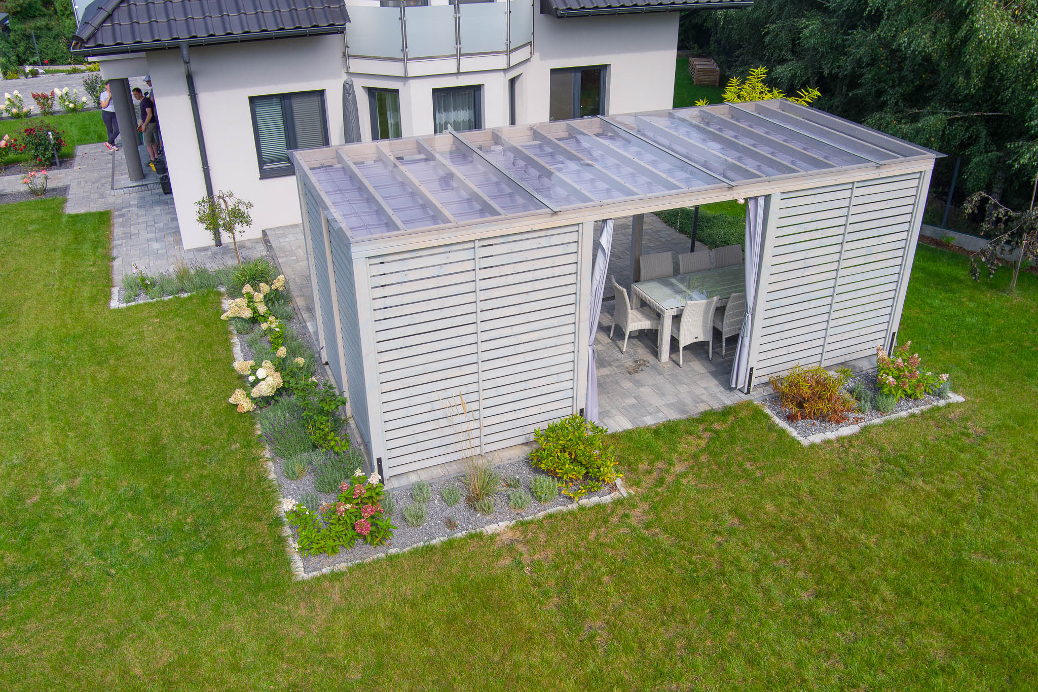 Zdjęcie z lotu ptaka przedstawia białą konstrukcję z drewnianych listew z przezroczystym dachem, wyposażoną w stół i krzesła, umieszczoną w ogrodzie przy nowoczesnym domu.