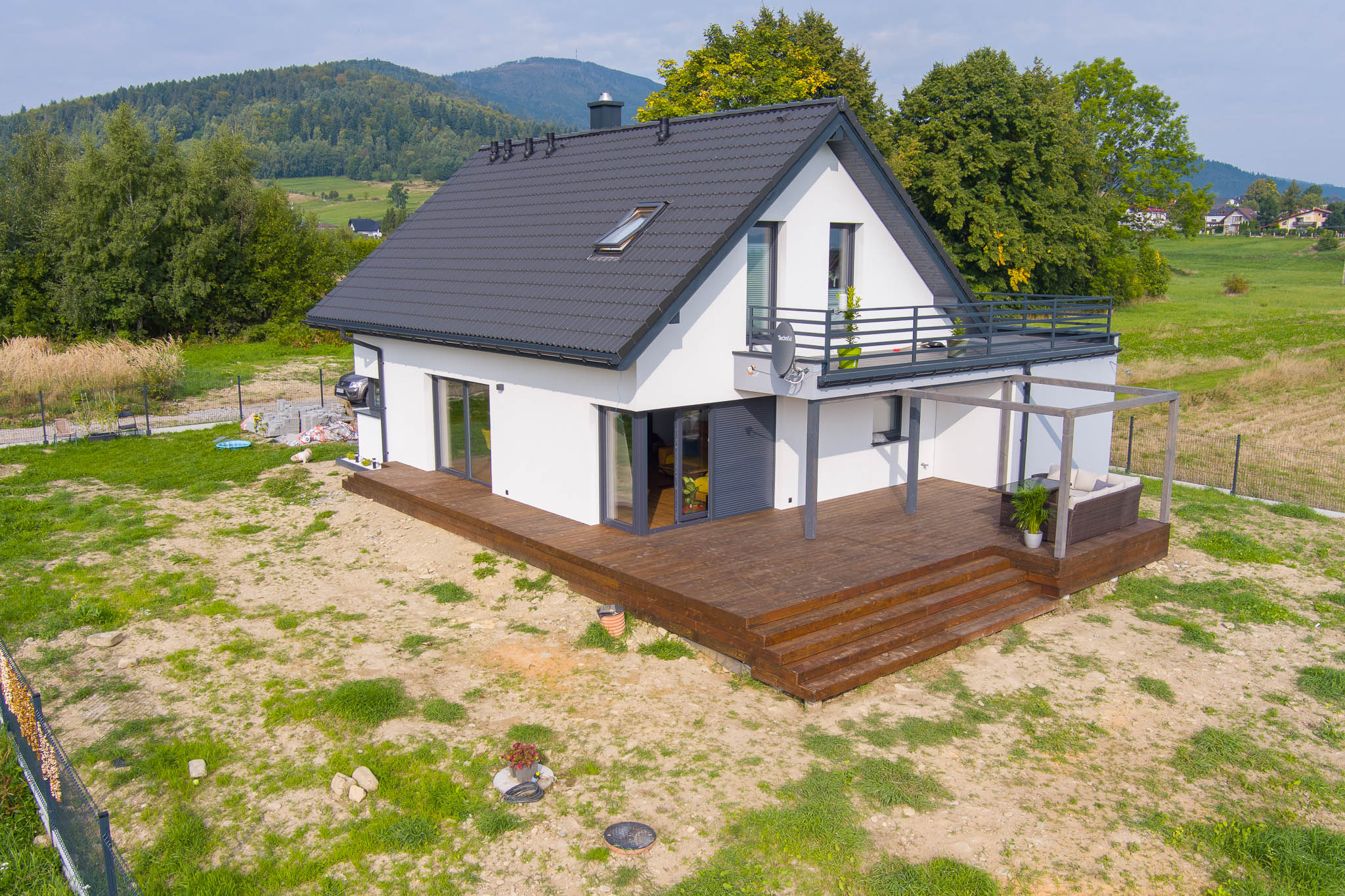 Nowoczesny dom z białymi ścianami i ciemnym dachem, z dużym tarasem z drewna i pergolą, widok z góry na tle zielonych wzgórz.