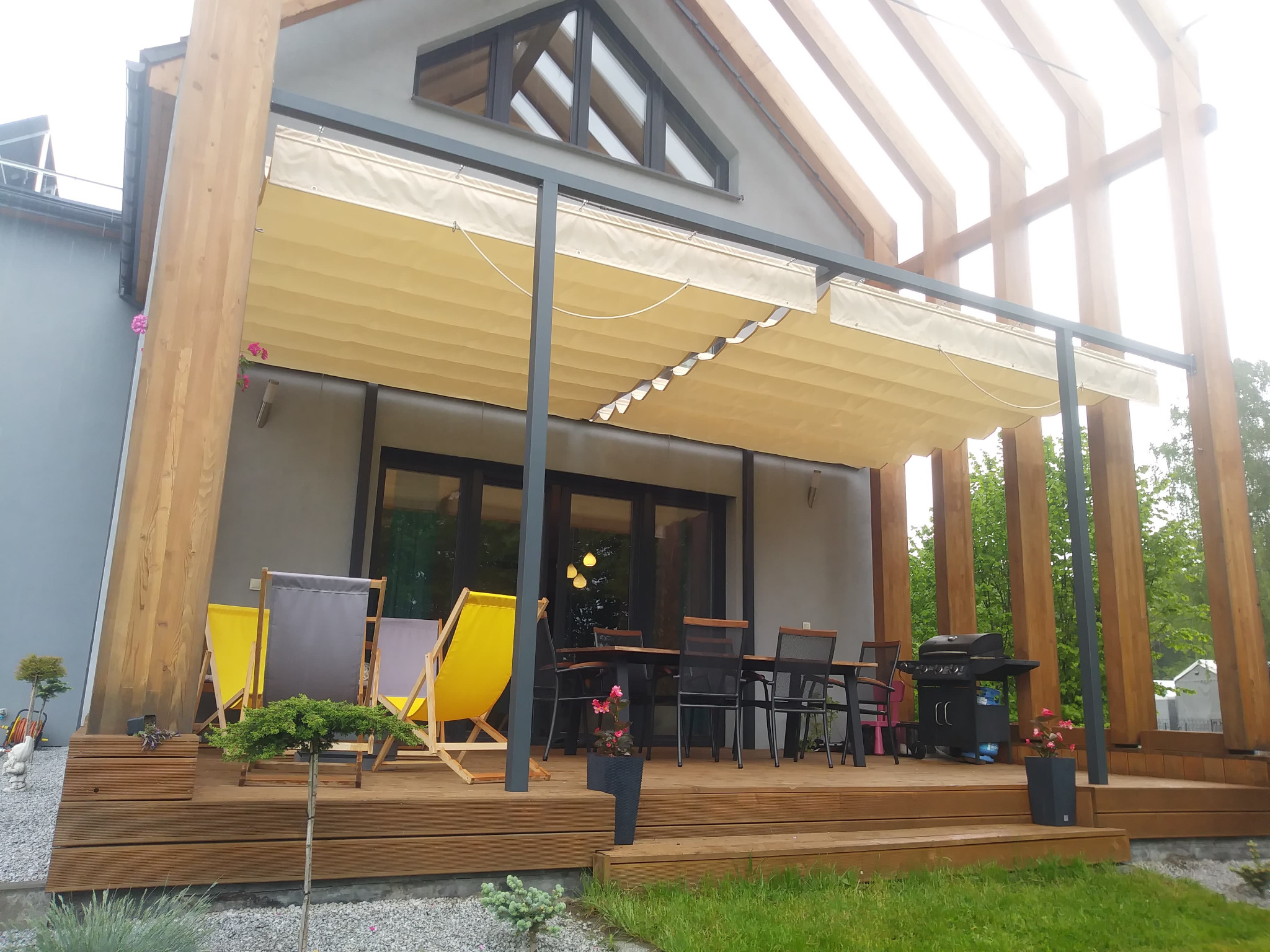 Zadaszony taras z drewnianą podłogą, meblami ogrodowymi i grillem, osłonięty beżową, zwijaną markizą, z widoczną konstrukcją drewnianą i zielenią w tle.