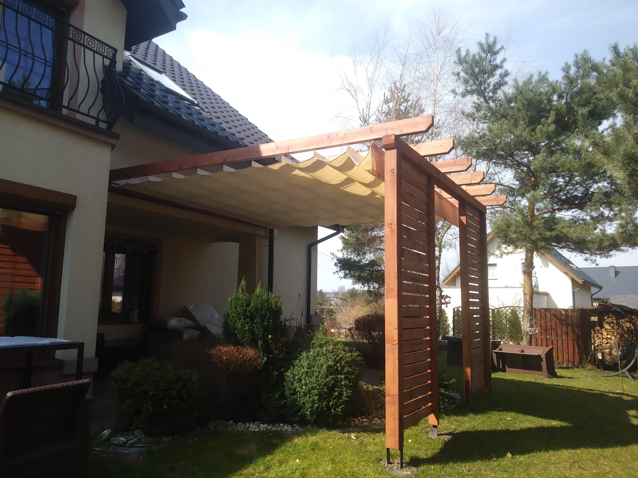 Drewniana konstrukcja z beżowym, falistym zadaszeniem przylegająca do domu, z widocznymi elementami ażurowej ścianki bocznej, na tle zielonego trawnika i iglastego drzewa.