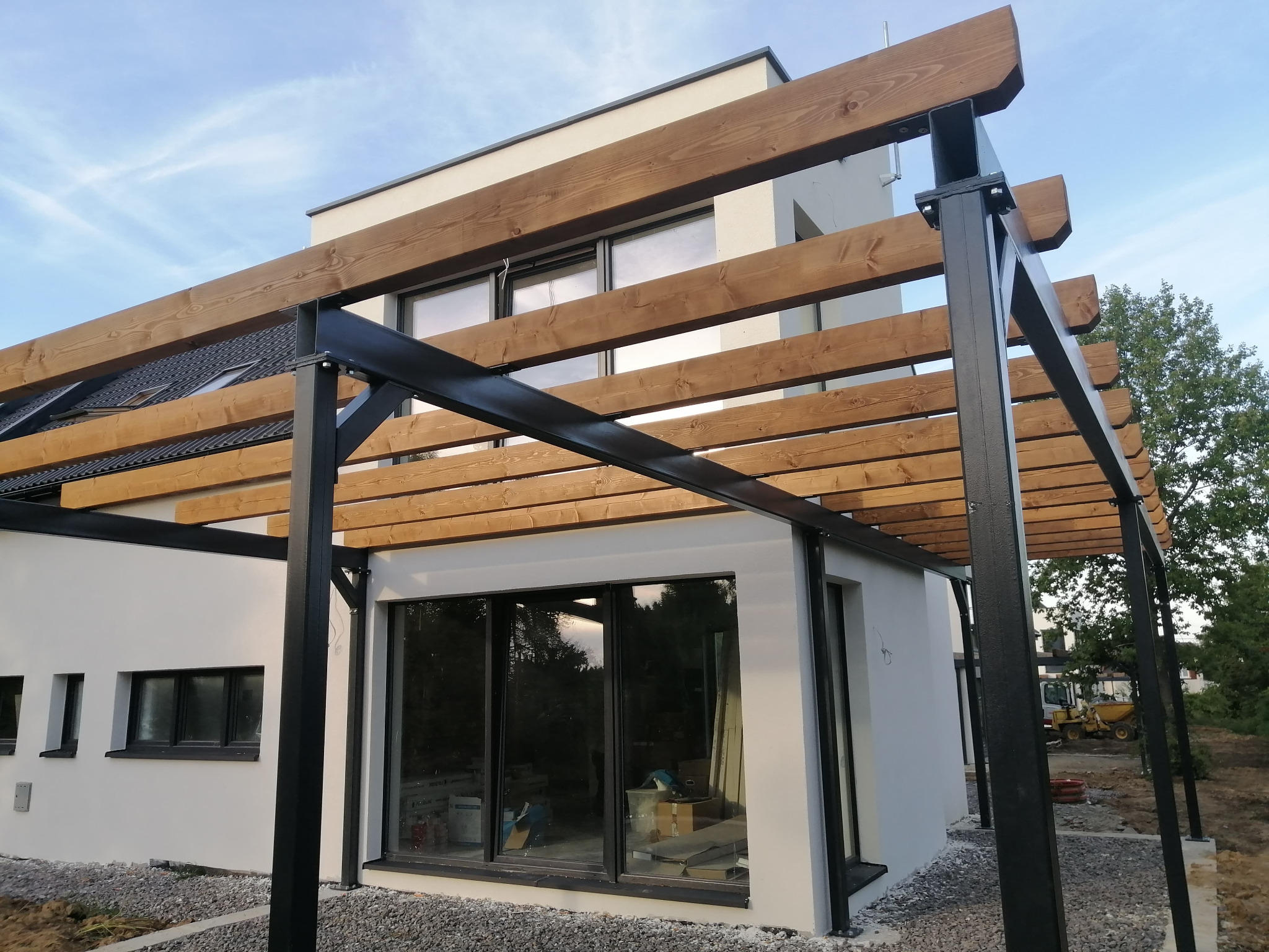 Nowoczesna konstrukcja z drewnianych belek i czarnych metalowych słupów przy budynku, widoczne okna i częściowo zagospodarowany teren.