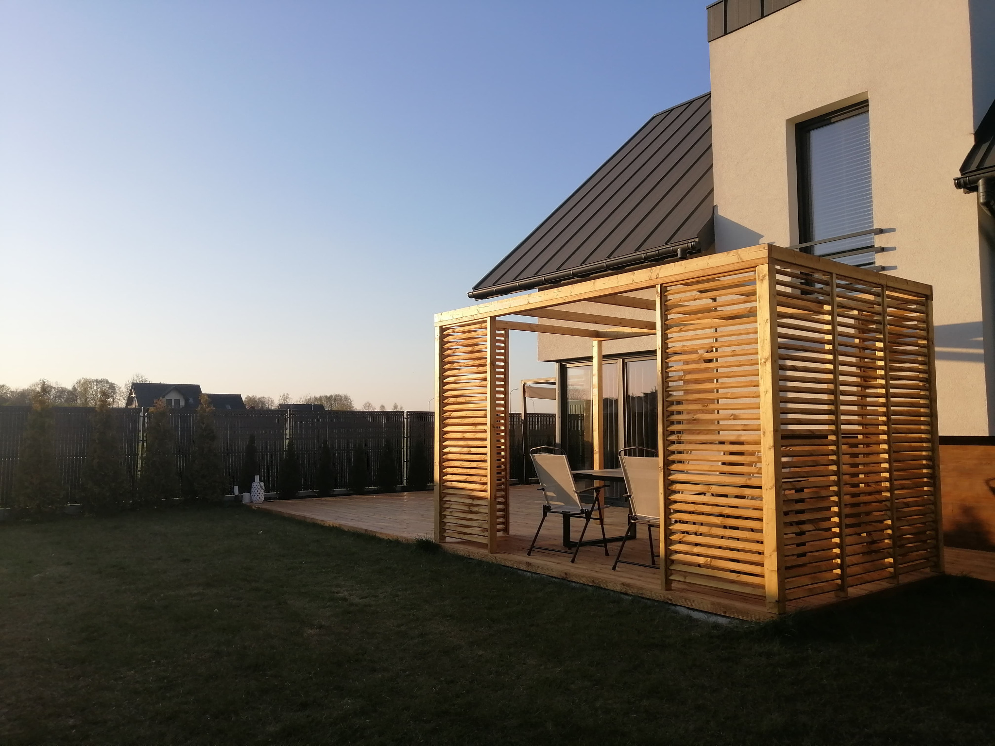 Nowoczesna konstrukcja z drewnianych listew tworząca zacienioną przestrzeń na tarasie, z widocznymi krzesłami i stołem, sąsiadująca z budynkiem o ciemnym dachu i ogrodzeniem w tle.