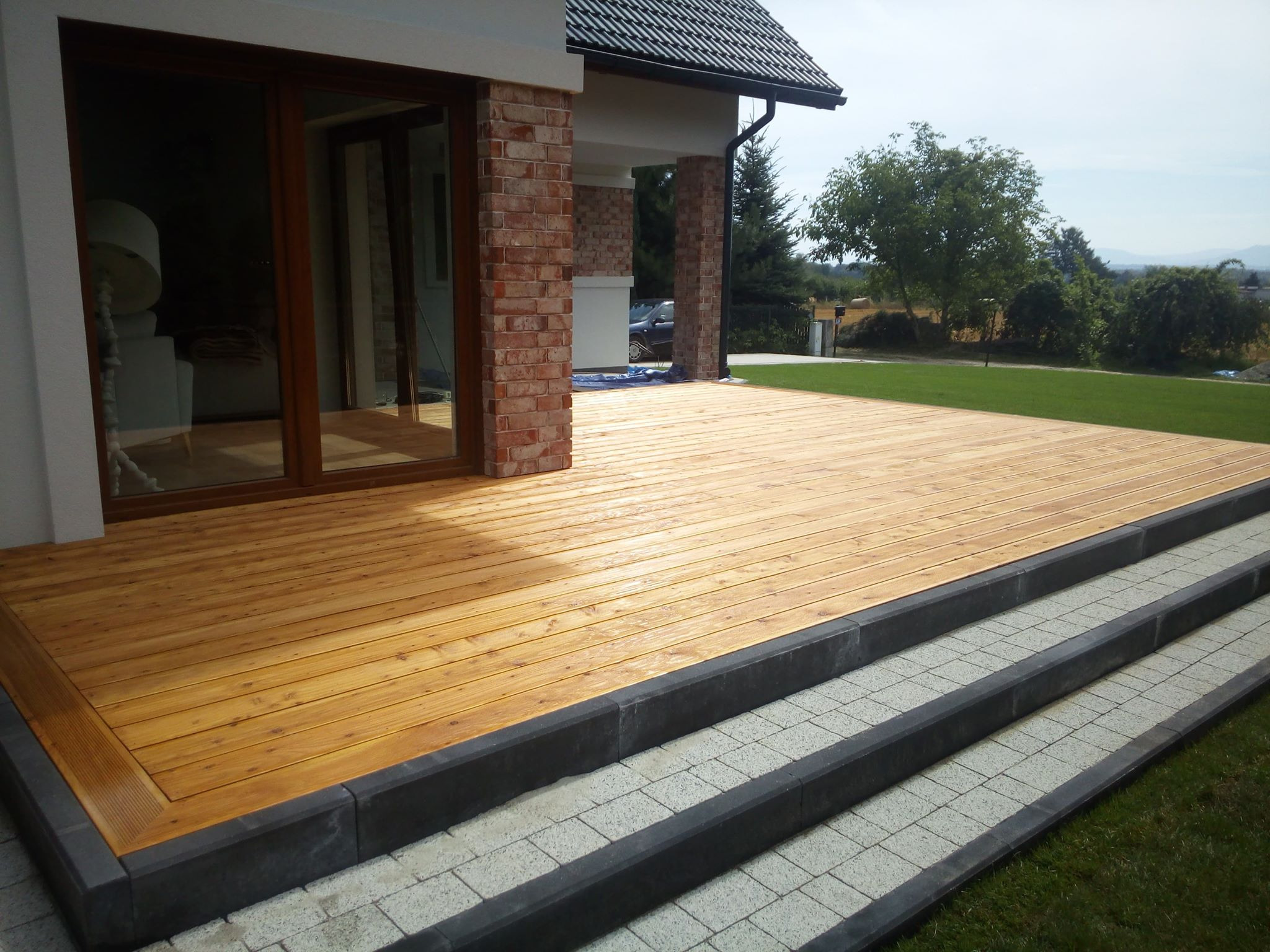 Jasna deska tarasowa z drewna na zewnątrz domu, z kamiennymi schodami i ceglanymi filarami, widok na zielony trawnik i drzewa w tle.