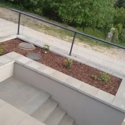 Schody z szarej kostki prowadzące do podwyższonego rabatu z roślinami, wykończonego korą i kamiennymi płytami, z widokiem na zieleń w tle.