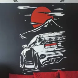 Czarno-biały mural na ścianie przedstawiający sportowy samochód na tle góry i czerwonego słońca, widoczny fragment kanapy z czerwonymi poduszkami.