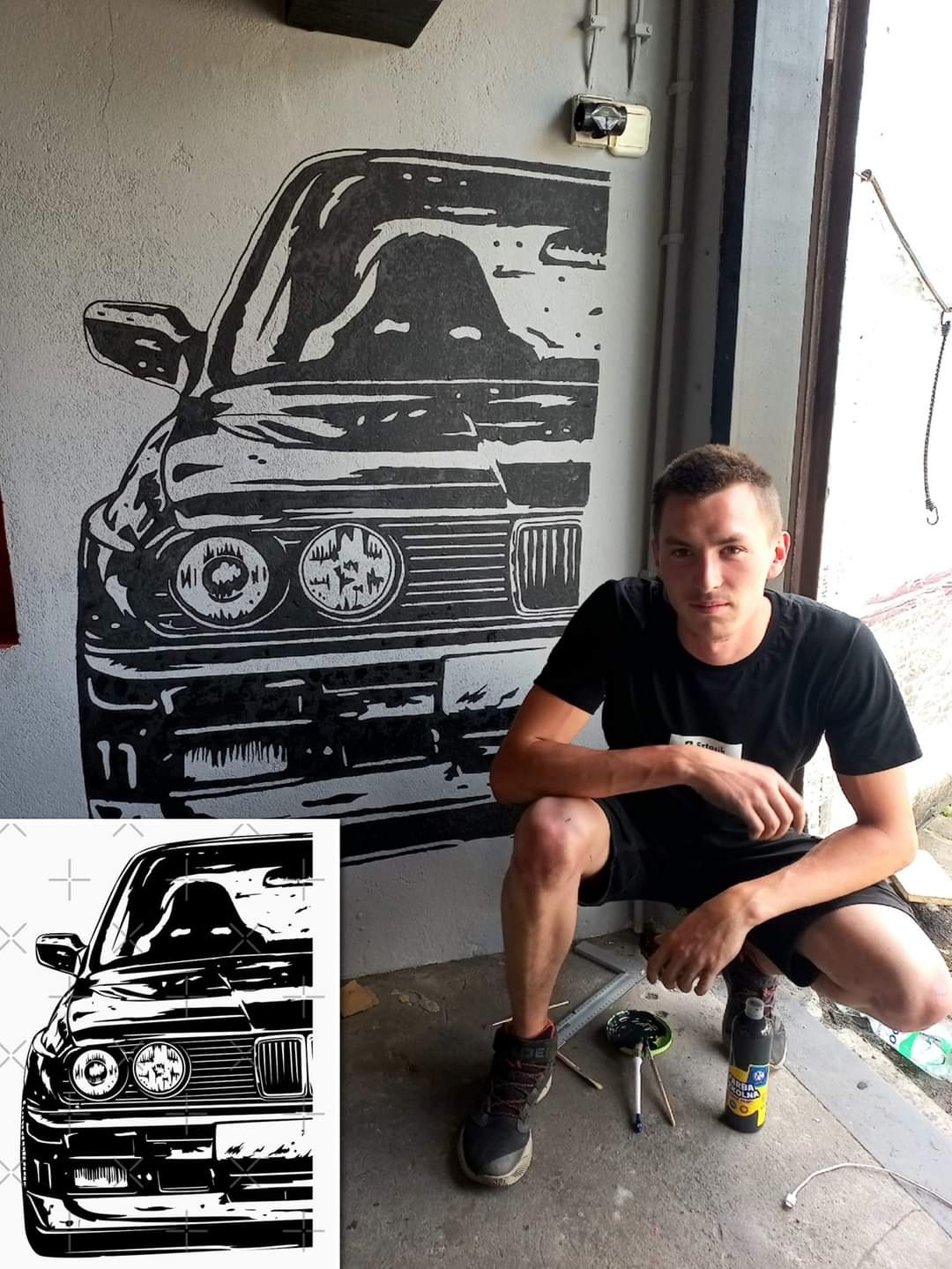 Mężczyzna kuca obok ściany z czarno-białym graffiti przedstawiającym fragment samochodu BMW E30, widoczne pędzle i farby.