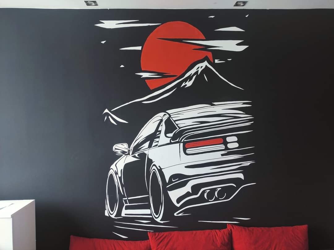 Czarno-biały mural na ścianie przedstawiający sportowy samochód na tle góry i czerwonego słońca, widoczny fragment kanapy z czerwonymi poduszkami.