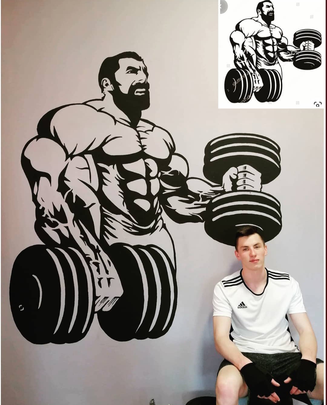 Czarno-biała grafika na ścianie przedstawiająca muskularnego mężczyznę z brodą podnoszącego hantle, obok siedzi młody mężczyzna w sportowym ubraniu.