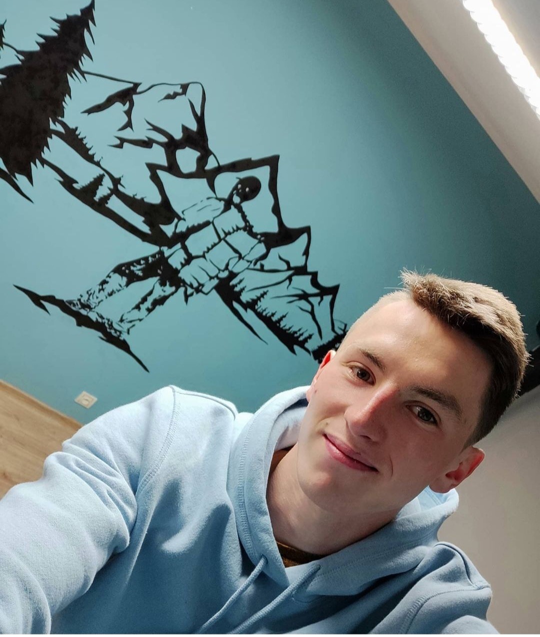 Uśmiechnięty mężczyzna w bluzie z kapturem, na suficie nad nim czarny rysunek gór i lasu na błękitnym tle.