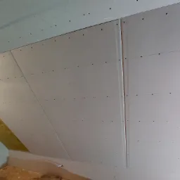 Skośna ściana poddasza pokryta płytami gipsowo-kartonowymi, widoczne ślady po wkrętach i fragment ocieplenia z wełny mineralnej, surowy stan wykończenia wnętrza.