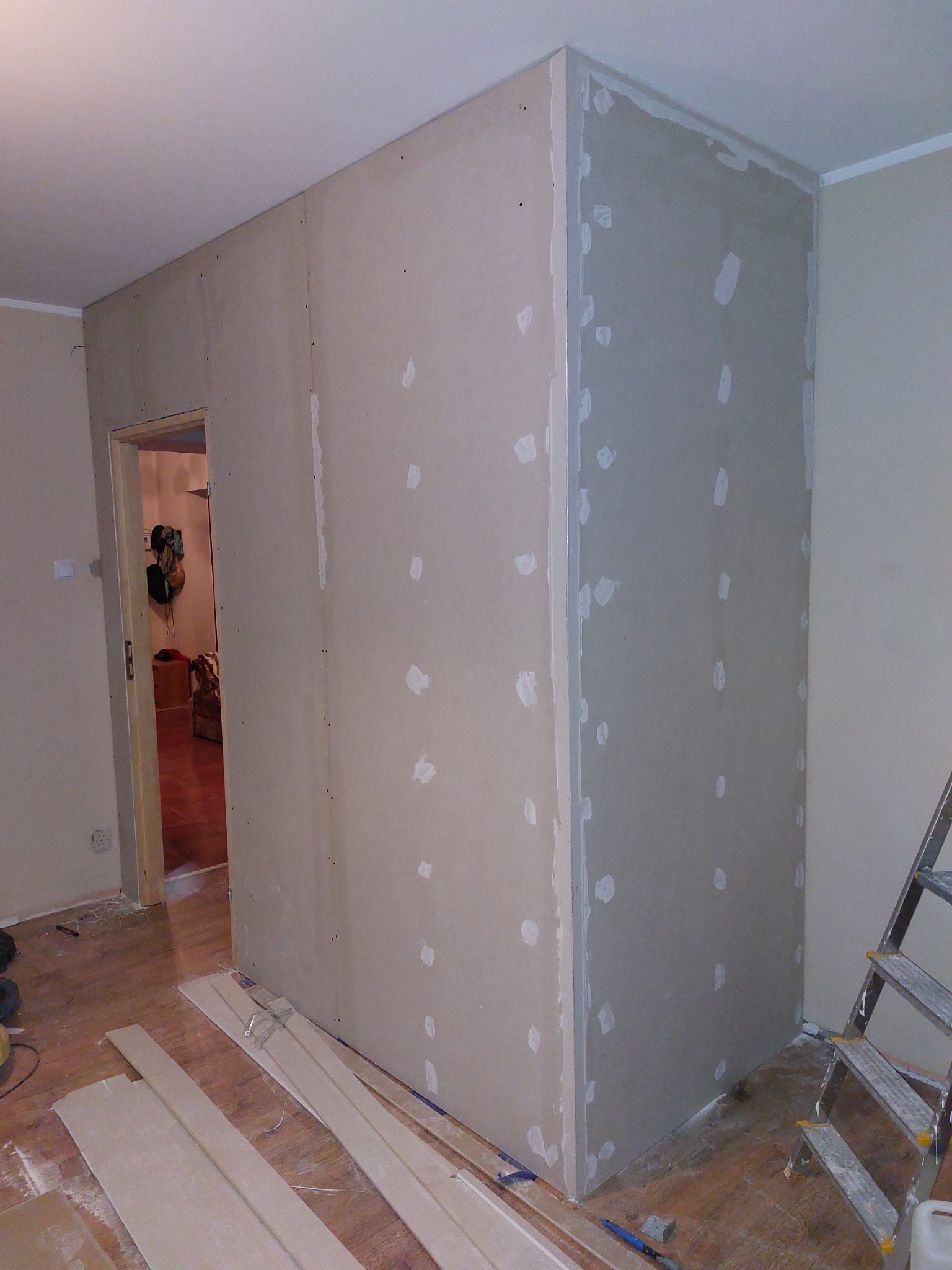 Konstrukcja z płyt karton-gips na metalowym stelażu, widoczne łączenia i śruby, przygotowana do szpachlowania, z fragmentem drabiny aluminiowej w prawym dolnym rogu, drewniana podłoga w tle.