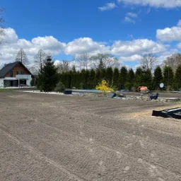 Przygotowany teren pod trawnik z widocznymi śladami po maszynach, w tle dom jednorodzinny z ogrodem, basen, panele słoneczne i betoniarka.