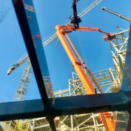 Widok z dołu na plac budowy z pomarańczową ładowarką teleskopową z koszem i dźwigiem, widziany przez metalową konstrukcję. W tle błękitne niebo.