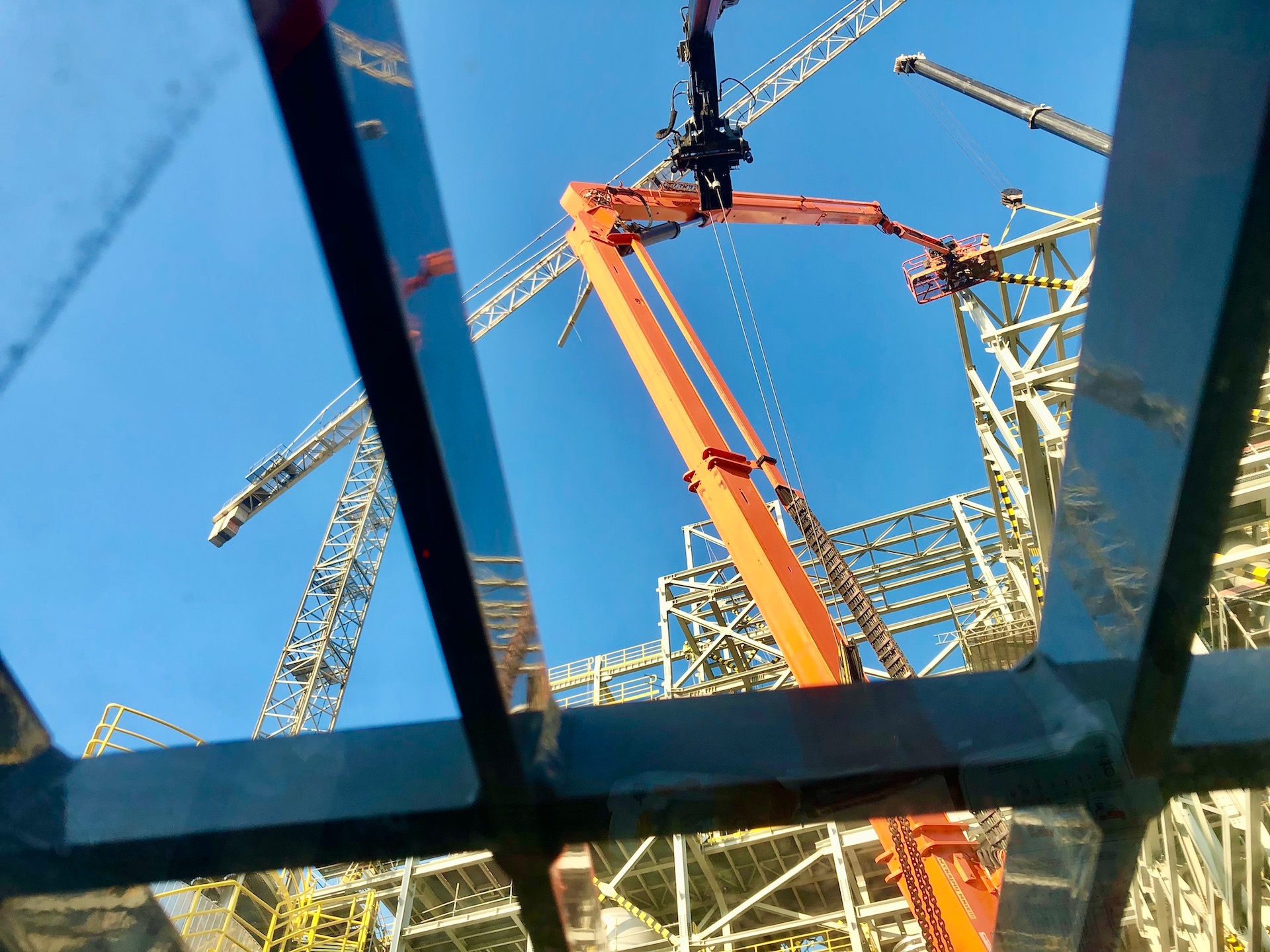 Widok z dołu na plac budowy z pomarańczową ładowarką teleskopową z koszem i dźwigiem, widziany przez metalową konstrukcję. W tle błękitne niebo.