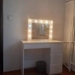 Biała toaletka z lustrem otoczonym żarówkami, ciemny kosz na śmieci pod blatem, parkiet na podłodze, widoczny fragment grzejnika na ścianie.