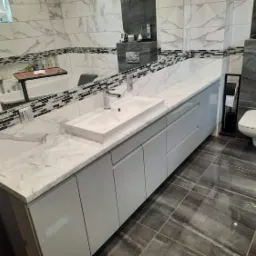 Nowoczesna łazienka z szarą marmurową płytką, białym blatem z umywalką i szarymi szafkami pod umywalką, widoczne lustro i toaleta.