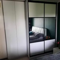 Biała szafa przesuwna z czarnymi ramami, częściowo lustrzana, odbijająca fragment sypialni z łóżkiem i kwiatami, obok szafa dwudrzwiowa, na podłodze narzędzia.