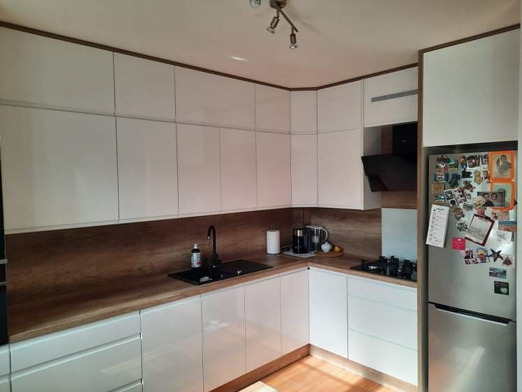 Biała kuchnia z połyskiem, drewniany blat i czarny zlew, widok na lodówkę oblepioną magnesami.