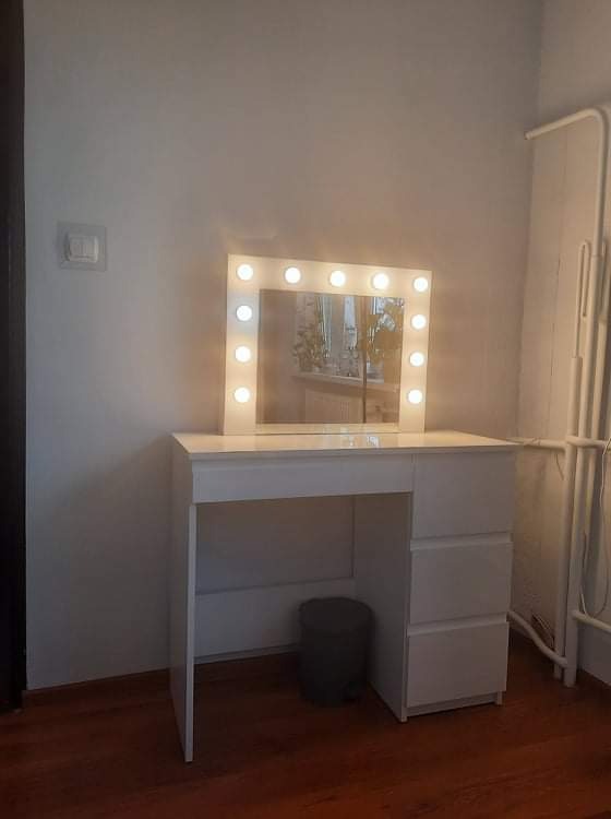 Biała toaletka z lustrem otoczonym żarówkami, ciemny kosz na śmieci pod blatem, parkiet na podłodze, widoczny fragment grzejnika na ścianie.