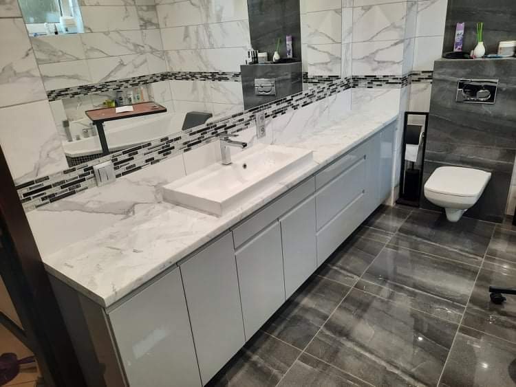 Nowoczesna łazienka z szarą marmurową płytką, białym blatem z umywalką i szarymi szafkami pod umywalką, widoczne lustro i toaleta.