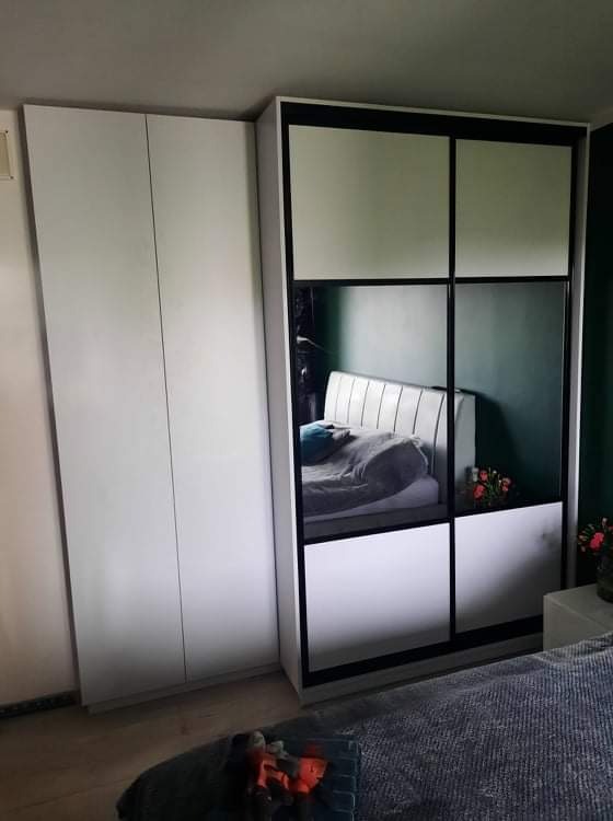 Biała szafa przesuwna z czarnymi ramami, częściowo lustrzana, odbijająca fragment sypialni z łóżkiem i kwiatami, obok szafa dwudrzwiowa, na podłodze narzędzia.