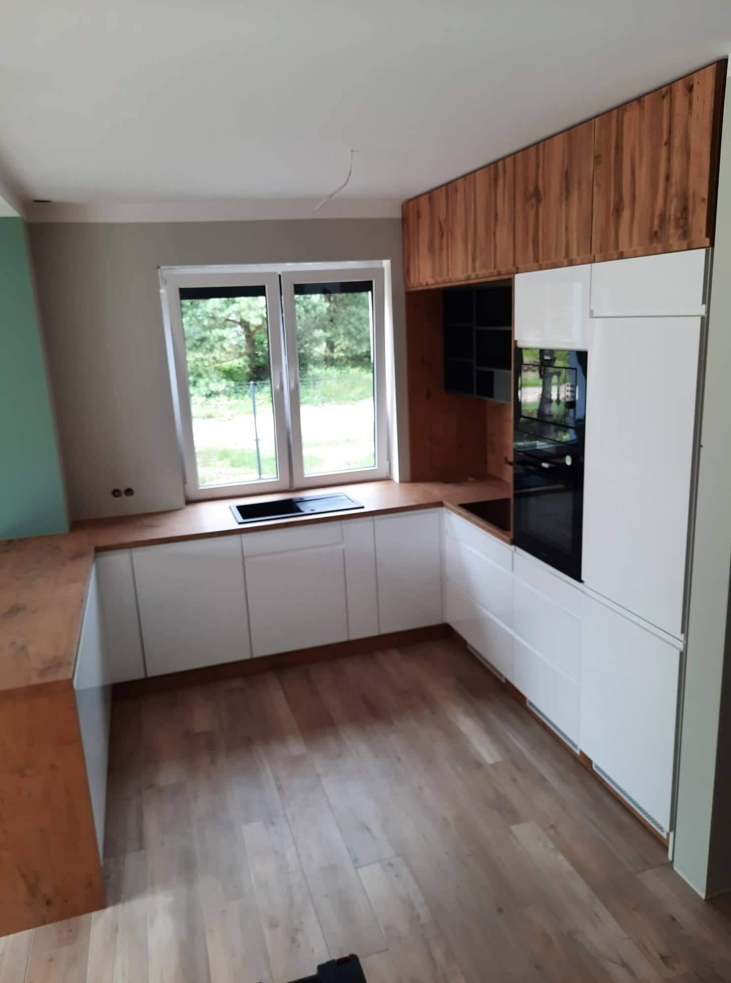 Nowoczesna kuchnia w kształcie litery L z białymi szafkami i drewnianym blatem, widok na okno i drewniane wykończenie górnych szafek.