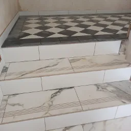 Wykończone schody z płytek imitujących marmur, z białymi podstopnicami i kontrastującą podłogą w szachownicę w tle, fragment ściany w stanie surowym.