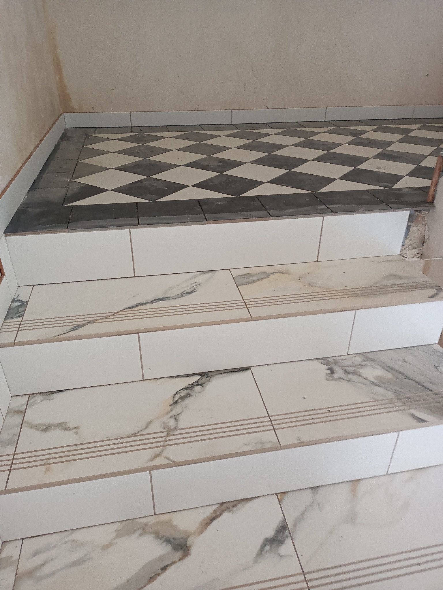 Wykończone schody z płytek imitujących marmur, z białymi podstopnicami i kontrastującą podłogą w szachownicę w tle, fragment ściany w stanie surowym.