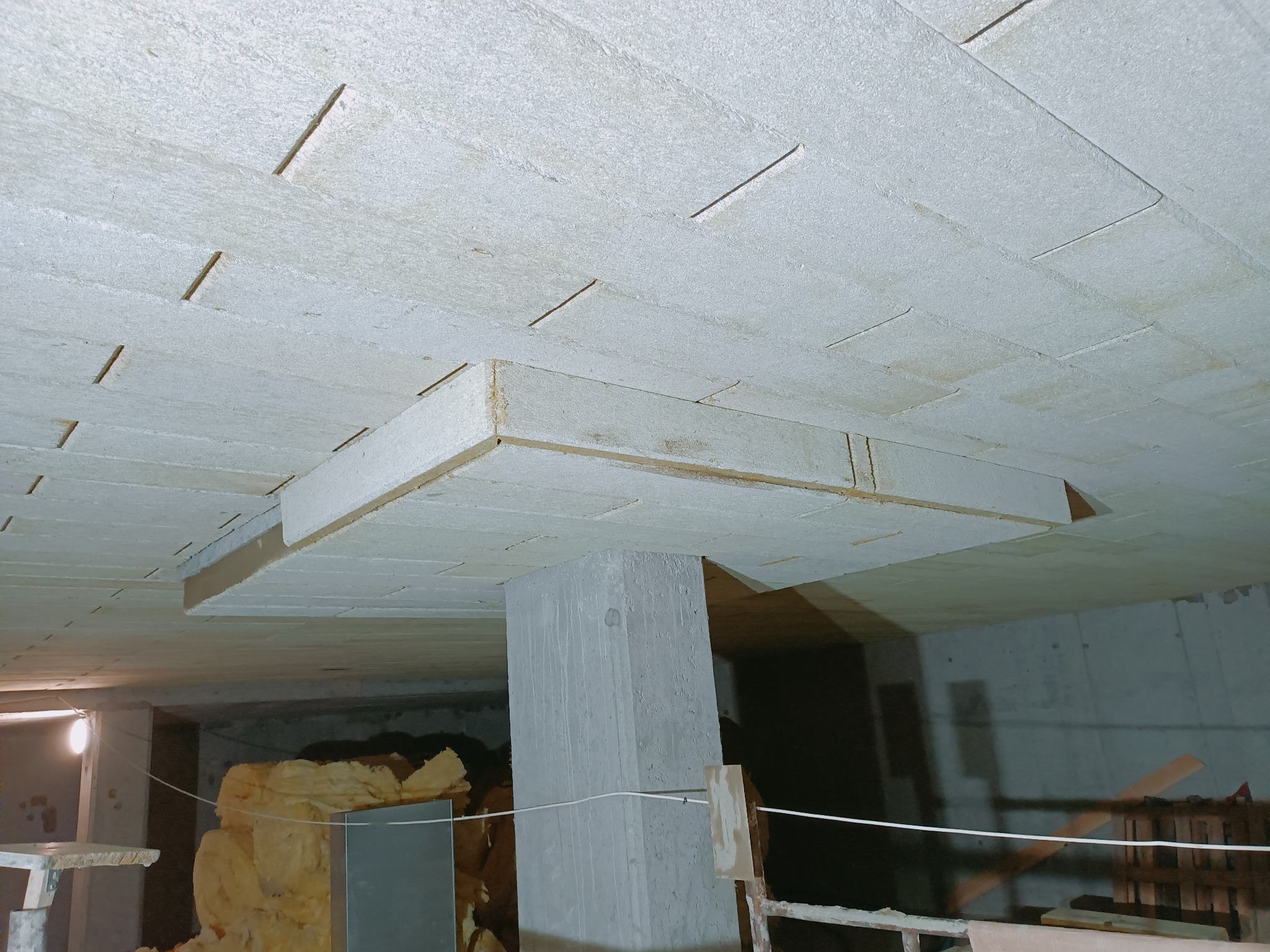 Sufit podwieszany z płyt w trakcie montażu, widoczne płyty z wyciętymi szczelinami, betonowy filar i fragment pomieszczenia w tle.