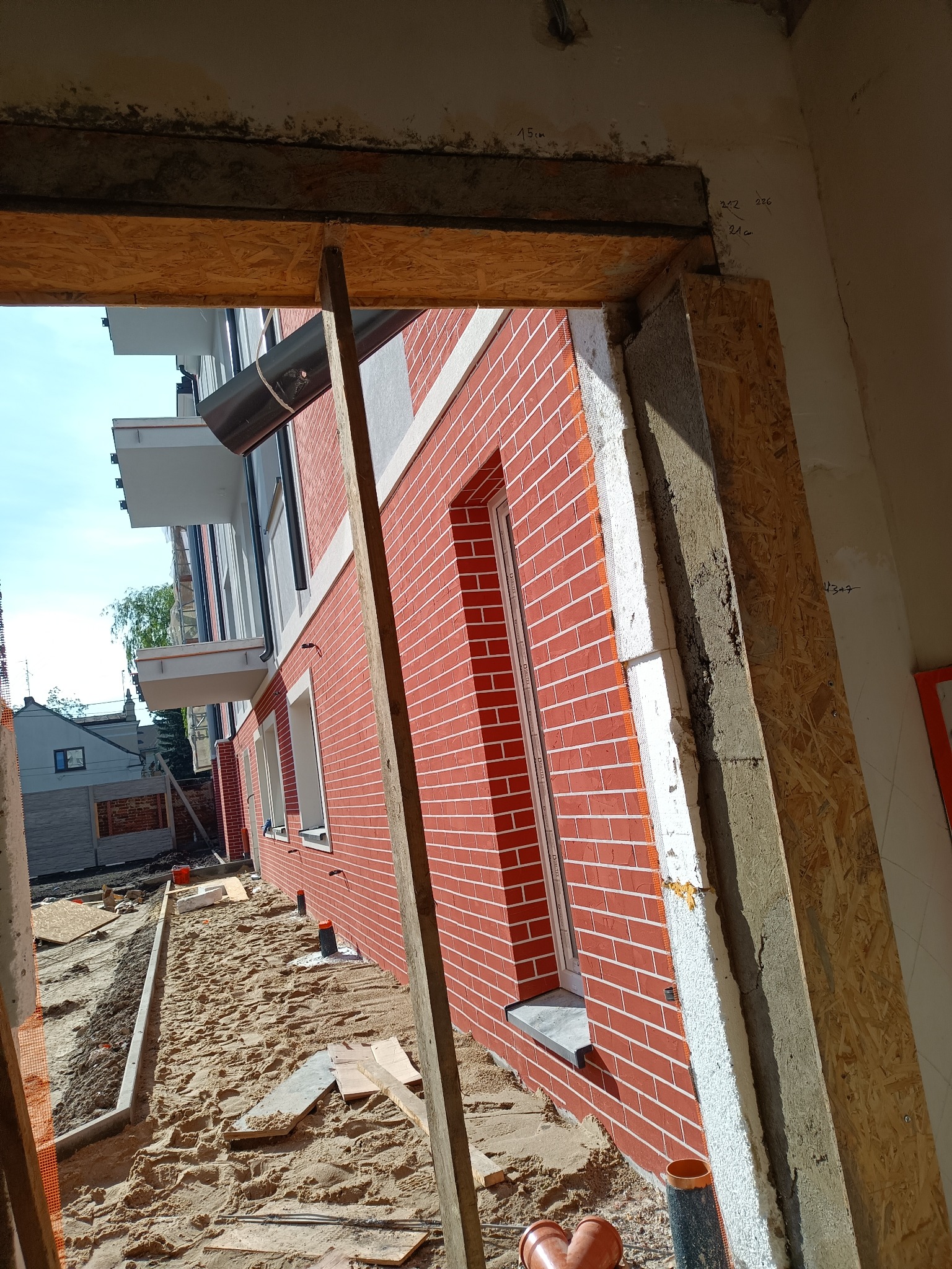 Budynek w trakcie ocieplania, widoczne warstwy izolacyjne, ceglana elewacja i otoczenie placu budowy z piaskiem.