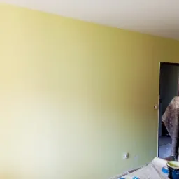 Świeżo pomalowana ściana na jasnożółty kolor w pokoju, z widocznymi śladami po taśmie malarskiej na podłodze i wiaderkiem z farbą w rogu.