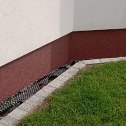 Fragment elewacji budynku z wykończeniem cokołu w kolorze ceglastym, obrzeżem z kostki brukowej i trawnikiem.