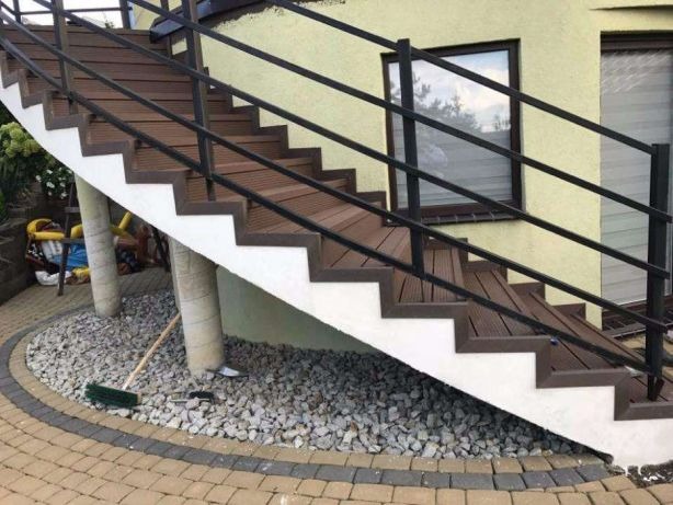 Zewnętrzne schody z desek kompozytowych z czarną metalową balustradą, prowadzące do budynku z oknem, otoczone kamieniami i kostką brukową.