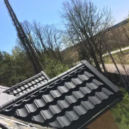 Fragment dachu pokrytego czarną blachodachówką modułową, widoczne elementy konstrukcyjne i obróbki blacharskie, w tle drzewa i błękitne niebo.