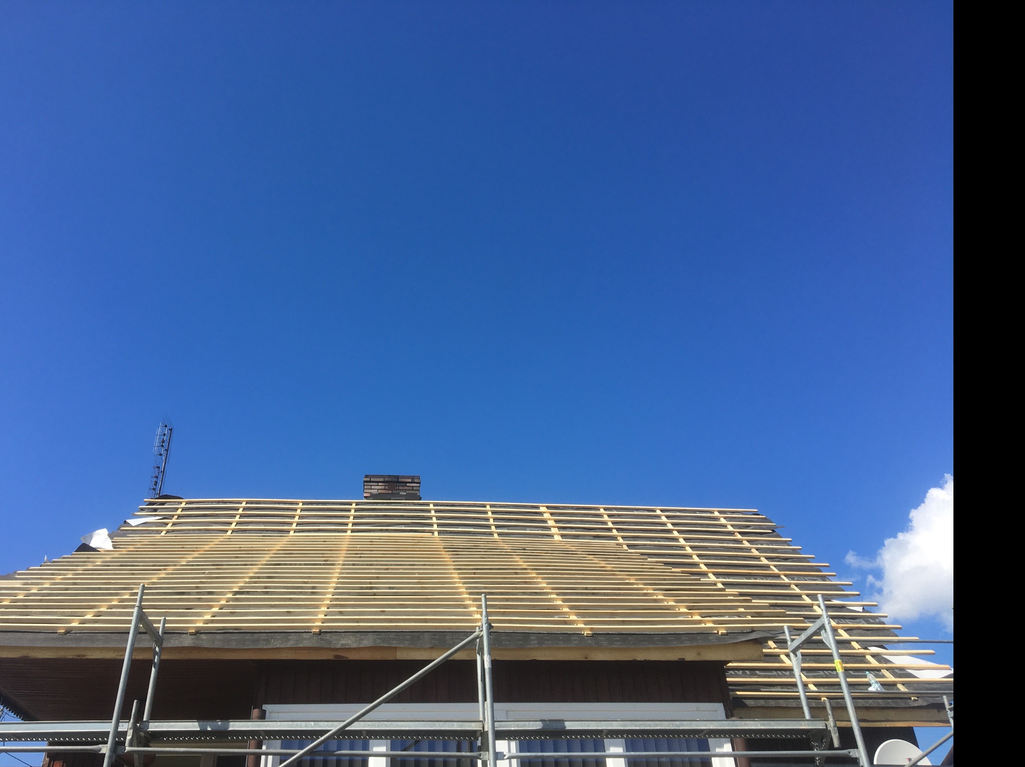 Widok z dołu na konstrukcję dachu z widocznymi łatami drewnianymi, rusztowaniem i kominem na tle błękitnego nieba.