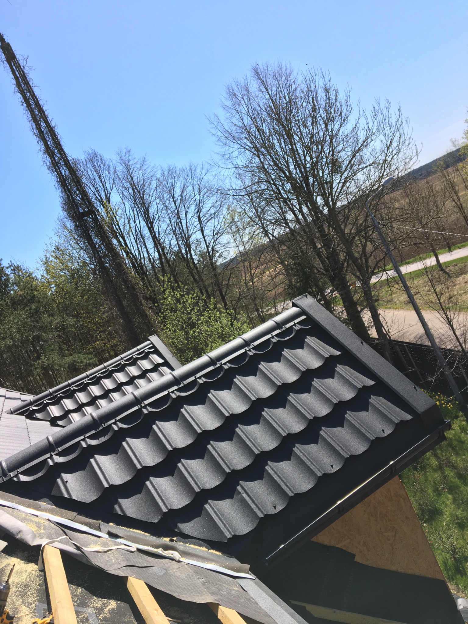 Fragment dachu pokrytego czarną blachodachówką modułową, widoczne elementy konstrukcyjne i obróbki blacharskie, w tle drzewa i błękitne niebo.