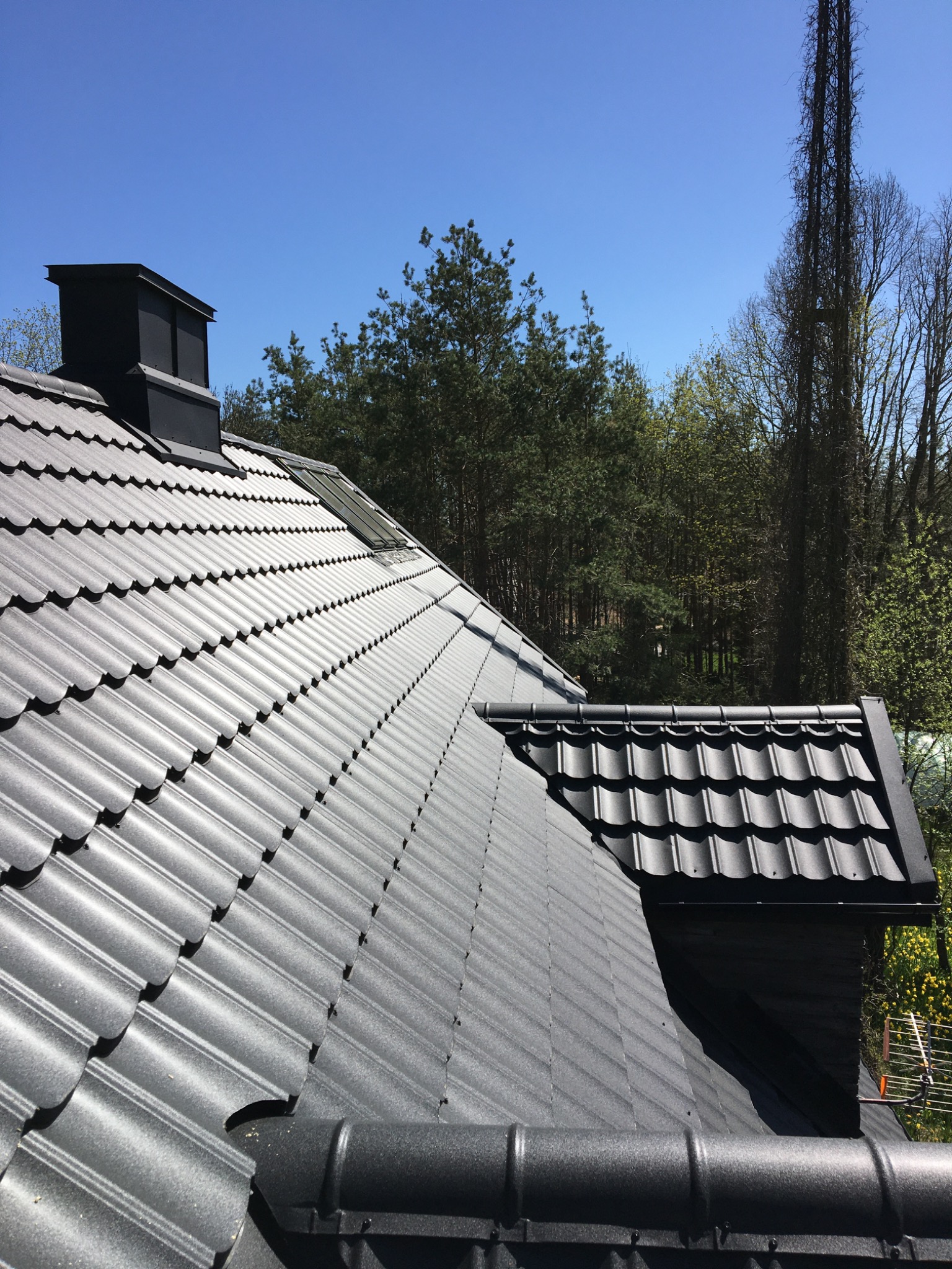 Szary dach z blachodachówki modułowej, widoczny komin, okno dachowe i rynna, w tle las i błękitne niebo.