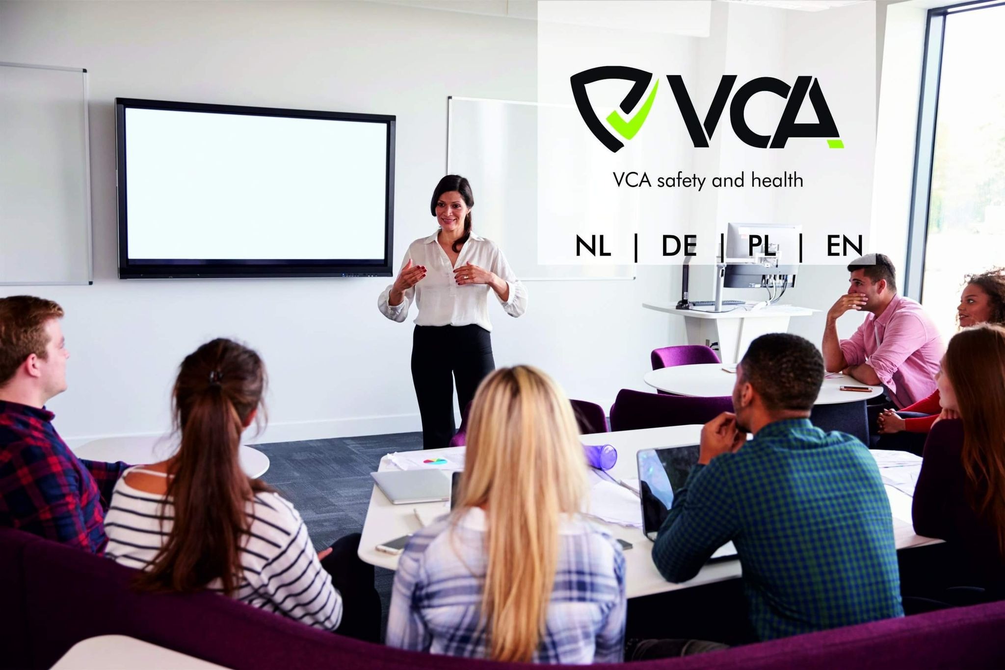 Prelegentka w jasnej koszuli gestykuluje podczas prezentacji przed grupą słuchaczy siedzących przy stolikach z laptopami; na ścianie logo VCA safety and health i ekran projekcyjny.
