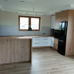 Nowoczesna kuchnia z wyspą, fronty szafek w kolorze białym i drewnopodobnym, czarna lodówka, srebrne płytki na ścianie, drewniane blaty i podłoga.