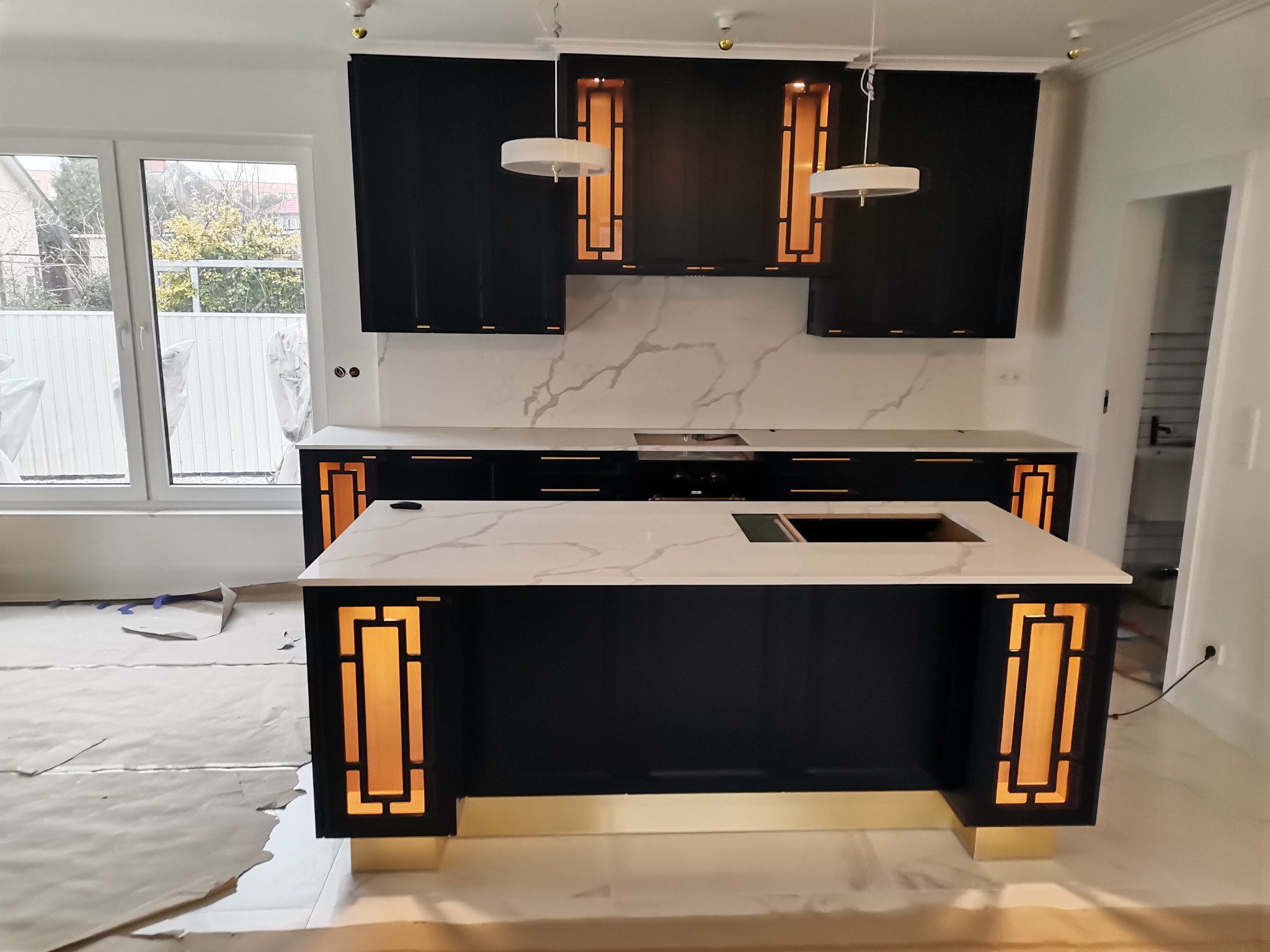 Nowoczesna kuchnia w trakcie montażu: czarne szafki z oświetleniem LED w złotych obudowach, biały blat z marmurowym wzorem, podłoga zabezpieczona folią ochronną.