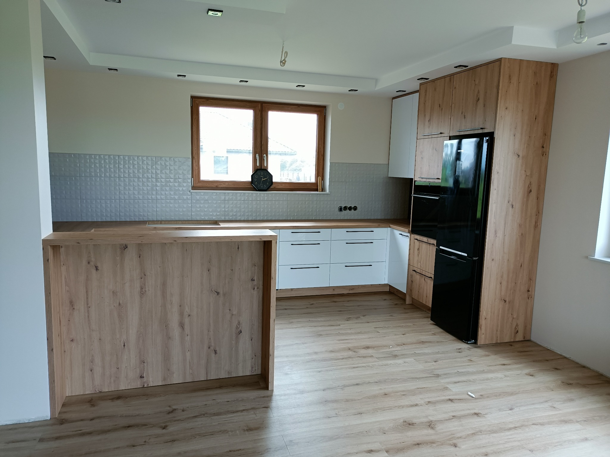 Nowoczesna kuchnia z wyspą, fronty szafek w kolorze białym i drewnopodobnym, czarna lodówka, srebrne płytki na ścianie, drewniane blaty i podłoga.
